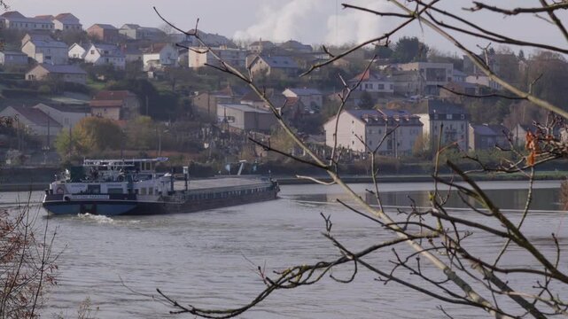Cargo barge crosses a calm river below a hillside town in Moselle, France.