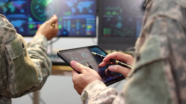 Military personnel in camouflage use a tablet and digital displays to review maps, data, and mission information in a modern command center.