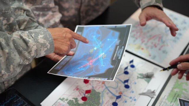Military personnel in camouflage study layered tactical maps and a blue digital navigation screen, pointing at routes and marked locations during a planning briefing.