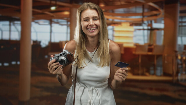 Woman holding a vintage camera and blue credit card, smiling while presenting the card in an airport terminal with seating and pillars; travel joy.
