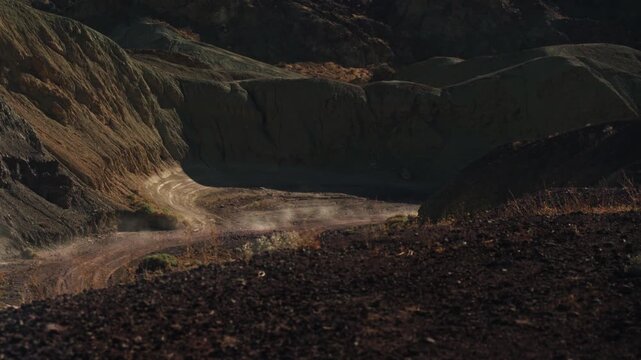 A dune buggy drives across a canyon floor, leaving a trail of dust behind it, shot from above