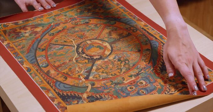 Traditional Nepalese Wheel of Life thangka is spread across a wooden table with the blue deity nearest the viewer and circular Buddhist scenes revealed before a hand rolls the red bordered scroll up