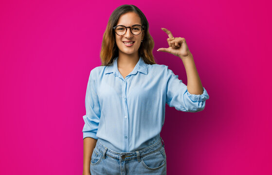Young woman hispanic clothes in studio, confident and smiling, hand and fingers showing small size sign &mdash; finger gesturing to measure, small sign concept