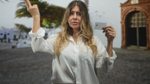 Woman holds creditcard and points finger to head on street with arched building and palm tree; frustration.