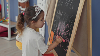 Girl drawing on chalkboard, hand holding pink chalk while sketching letters in preschool classroom  curiosity learning imagination. © Krakenimages.com
