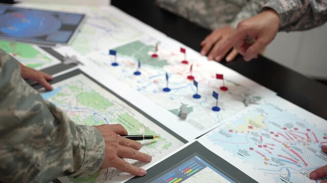 Military personnel in camouflage study maps with colored markers and digital tablets during a strategic planning meeting, showing teamwork, navigation and mission preparation.