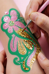 Naklejka premium Artist applies gold sequins to glitter flower tattoo on child arm. Pro makes temporary body art with pink sparkles, green patterns. Creative glitter makeup for kids at festive party birthday event