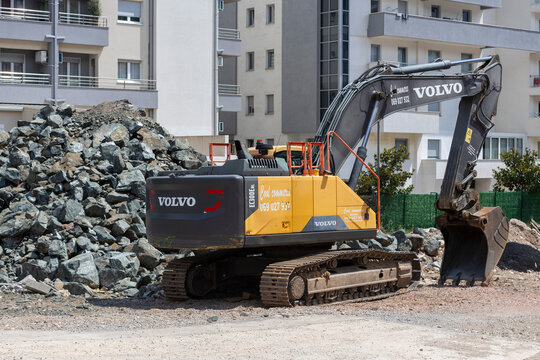 July 1, 2026, Budva, Montenegro, Excavator Working on Construction Site with Large Rock Pile and Modern Buildings in Background