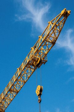 Large yellow construction crane jib extends toward blue sky with white clouds. Heavy industrial machinery uses steel lattice boom, lifting hook for building projects. Massive metal equipment works at