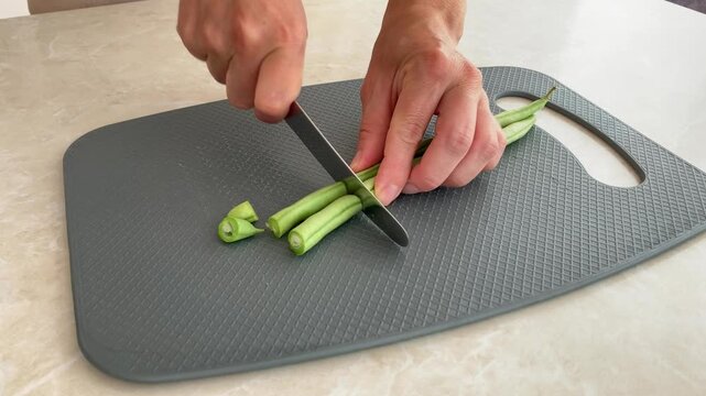 The woman is cutting fresh green beans with a metal knife on a gray cutting board; the cook is preparing seasonal vegetables for a home‑cooked dish; fresh harvest and healthy eating