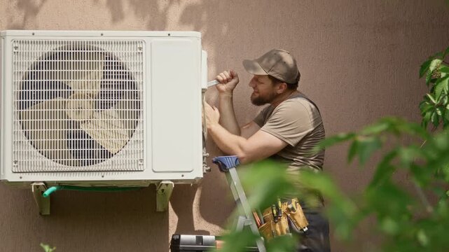 Caucasian young adult technician mounting outdoor air conditioner unit on wall with wrench. Footage fits HVAC maintenance campaigns, contractor commercials, and home service promos