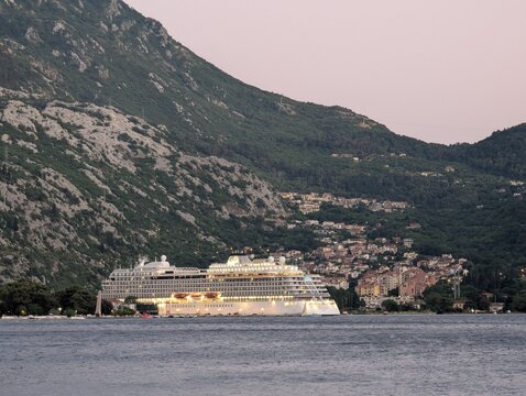 July 3, 2026, Kotor, Montenegro, Cruise ship docked near a mountainous coastline with illuminated town at dusk and calm water