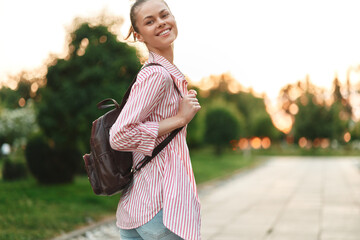 Fototapeta premium Young woman with a striped shirt and backpack smiles while walking along a sunlit path at dusk, conveying a carefree, hopeful mood and a sense of spontaneous everyday adventure.