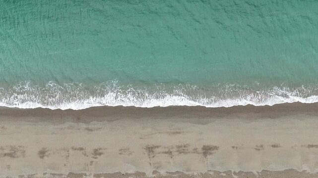 Top down drone footage of turquoise sea waves gently breaking along a sandy beach, creating a clean natural coastal pattern from above.
