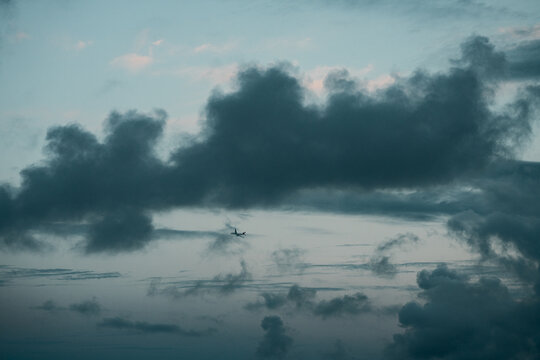 An airplane crosses the sky beneath a broad cloud. The small silhouette sits near the center below a gray mass with pale blue gaps around it.