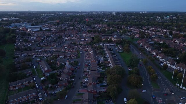Aerial drone flight over a typical residential neighborhood in England during blue hour after sunset, showing rows of British houses, suburban streets, moving cars, green spaces and the surrounding ur