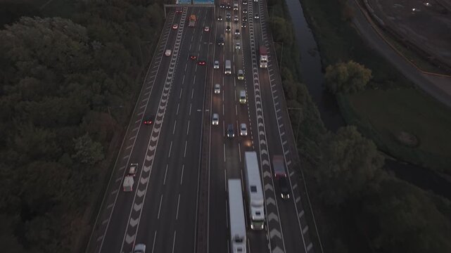 Drone flies forward above a busy motorway in England toward oncoming traffic, passing over cars, vans and large trucks. Aerial view of vehicles travelling along multiple lanes at dusk, illustrating co