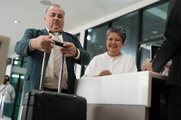 A senior traveller at the check in desk leans on his suitcase handle at the counter in the departure hall of a modern airport, good for senior and business travel themes © ultramansk