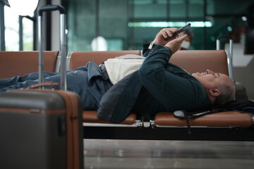 A weary traveller resting on the terminal seating lies across the waiting room bench with his hands folded in the waiting area of a modern terminal, good for disruption and fatigue themes © ultramansk