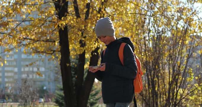 Schoolboy with backpack using smartphone in autumn park. Independent student spends time outdoors while staying connected, representing modern childhood, mobility and everyday school routine.