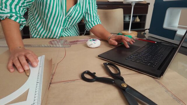 Woman fashion designer working on a laptop while drawing patterns on brown paper in her studio