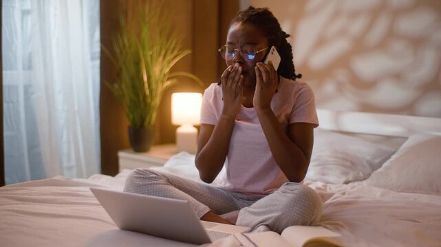 Tired African American business student woman yawning multitasking on bed home bedroom late evening sleepy female freelancer using laptop mobile phone call taking notes overworked girl working night