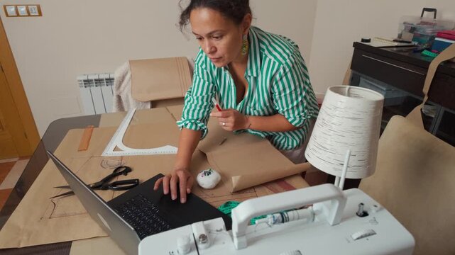 A woman tailors clothes at home, using a laptop, cutting paper patterns, and designing with a sewing machine.