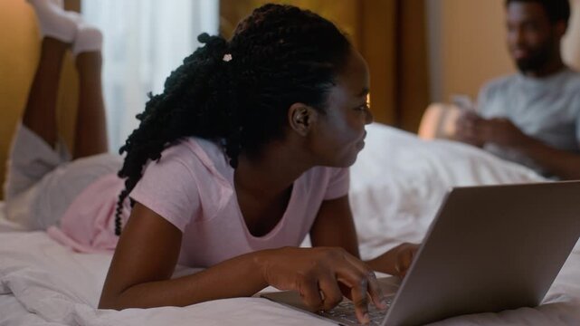 African American happy couple using laptop and mobile phone at night in bedroom girlfriend typing computer boyfriend chatting smartphone family weekend online shopping technology gadget addiction home