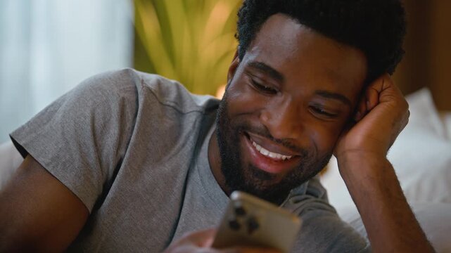 Close up laughing happy African American man holding mobile phone lying in bed scrolling social media at home bedroom late night male guy user cellphone online chatting gadget addiction bedtime indoor