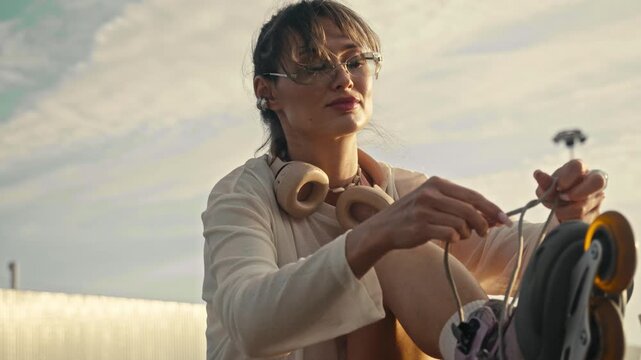 Young woman tightly pulls laces on inline skates at sunset. Beautiful girl wearing glasses prepares for active outdoor workout near water bottle in urban sports park during warm golden hour.
