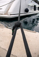 Fototapeta premium Black braided mooring rope knotted on quay with luxury yacht stern in blurred background.