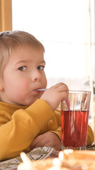 Child boy with blonde hair baby drinking lemonade in cafe. 3 years old kid drinks red juice from glasses through straws. Boy toddler in a restaurant. Closeup natural emotion vintage portrait. © Marina Demidiuk