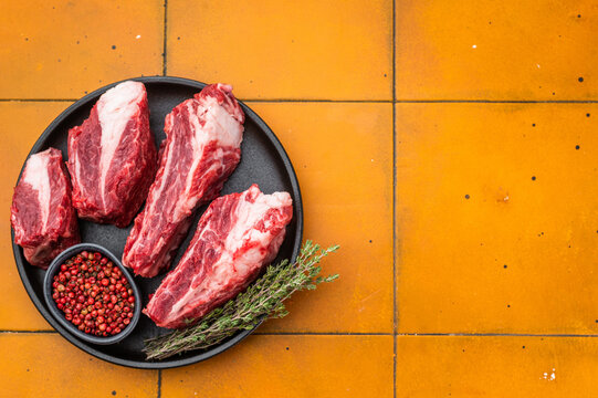 Raw beef short ribs chunks with seasoning and herbs on black plate. Vibrant yellow background with copy space for recipe concepts.