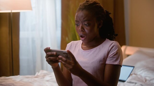 Shocked African American woman watching video on smartphone at home bedroom night upset disappointed ethnic female girl using cellphone negative reaction mobile phone gaming failure losing game indoor