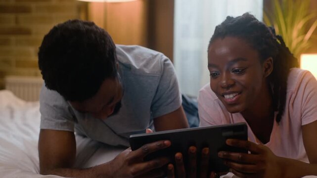 Happy African American couple laughing together using tablet at home bedroom evening married family watching funny online movie wife husband digital device comedy amusement relaxation relationship