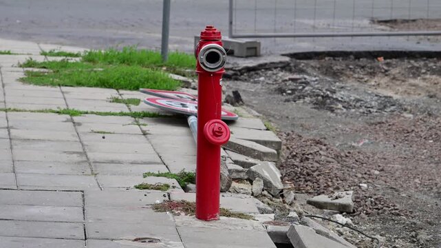 A bright red fire hydrant stands on a damaged concrete pavement next to an active street roadwork zone and a fallen traffic sign.
