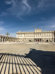 Scenic view of the Royal Palace of Madrid courtyard under a bright summer sky, showcasing baroque architecture, historic stone arches, and fence shadows. © Dragoș Asaftei