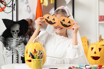 Fototapeta premium Halloween celebration and childhood creativity, happy girl wearing a bridal veil holding paper pumpkin masks over her eyes at home during autumn