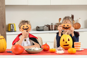 Fototapeta premium Halloween celebration and family fun, children in costumes holding paper pumpkin masks while sitting at a kitchen table with carved jack-o-lanterns