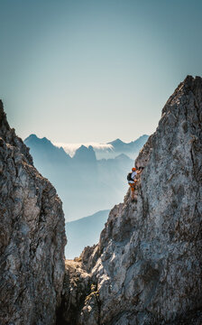 Solo male climber ascending a vertical rock wall on a via ferrata in the Austrian Alps near Innsbruck, Tyrol, with rugged cliffs and distant mountain peaks creating a dramatic alpine scene.