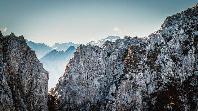 Solo male climber ascending a vertical rock wall on a via ferrata in the Austrian Alps near Innsbruck, Tyrol, with rugged cliffs and distant mountain peaks creating a dramatic alpine scene.