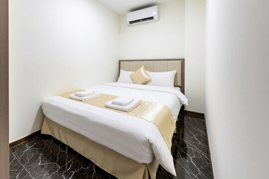 Small modern hotel room with double bed, white bedding. Gold pillows, bed runner add luxury touch to bedroom interior. Air conditioner mounted on wall above bed. Dark marble floor creates elegant