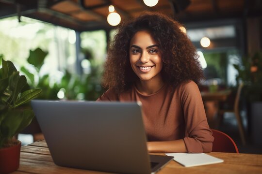 Young woman with curly hair confidently smiling at the camera, actively working on a laptop in a cozy cafe, embracing remote work, entrepreneurship, and flexible professional life