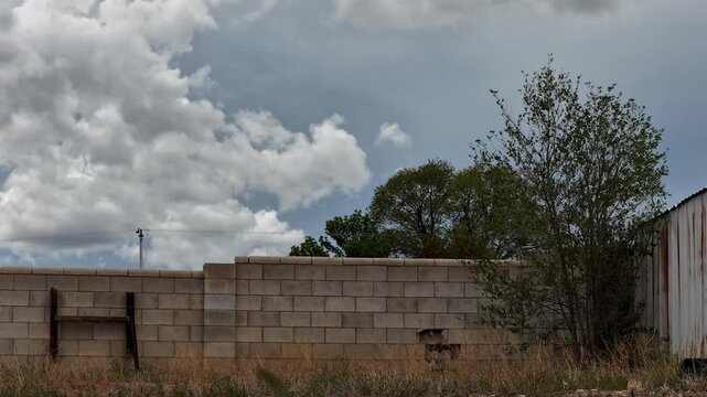 4K steady ground time lapse of massive fog and thick cumulus clouds rising behind a residential cinderblock wall. Dry grass, trees, and a metal shed frame the dynamic, shifting overcast sky.