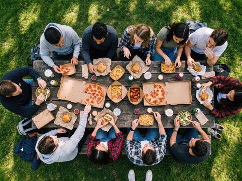 Friends sharing casual takeout food at an outdoor wooden picnic table, overhead view. Many people gather around a long weathered table covered with pizza, burgers, french fries, chicken wings, salads,