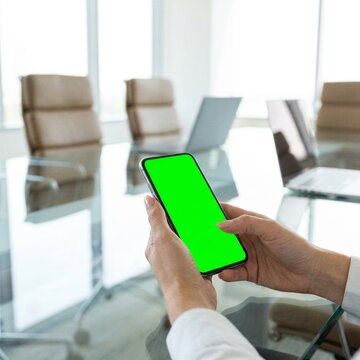 Business person holding a smartphone with a green screen in a modern office boardroom.