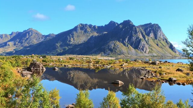 Picturesque landscape along Fv815 near Stamsund on Vestvagoya with a tranquil bay, impressive mountain range and nearly perfect reflection in the water beneath blue sky.