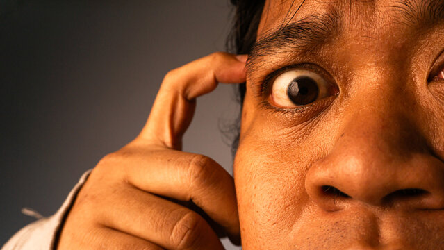 Detailed extreme close up portrait featuring young Asian male holding index finger against temple while staring intensely. Cropped composition focuses heavily on one wide eye, displaying