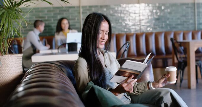 Mid adult Asian woman on tufted sofa in cafe, reading brochure, flipping pages and sipping coffee