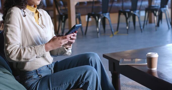 Mid-adult Asian woman receiving alert, tapping phone, smiling on sofa in cafe wearing beige shirt